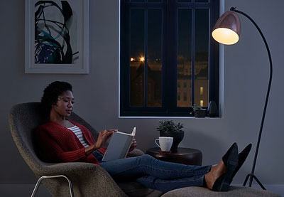 A woman sitting in a sofa reading a book. In front is a floor lamp with a smart bulb turned on   